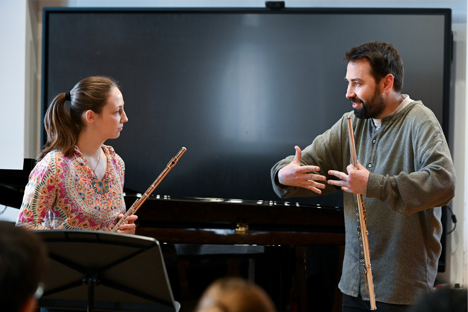 A male professor, holding a flute, teaching a female flautist on breathing, with a stand in between them and audience members looking at them in the foreground.